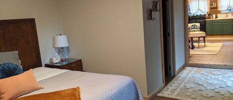 Un dormitorio con cama, mesita de noche, lámpara y vista a la cocina.