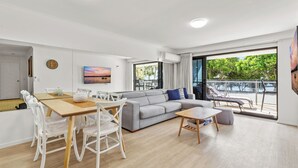 A modern living room with a dining area, grey sofa, and a large window with a view of the outdoors. - Spinnaker Apt 6- Mooloolaba Beach  (Mooloolaba)