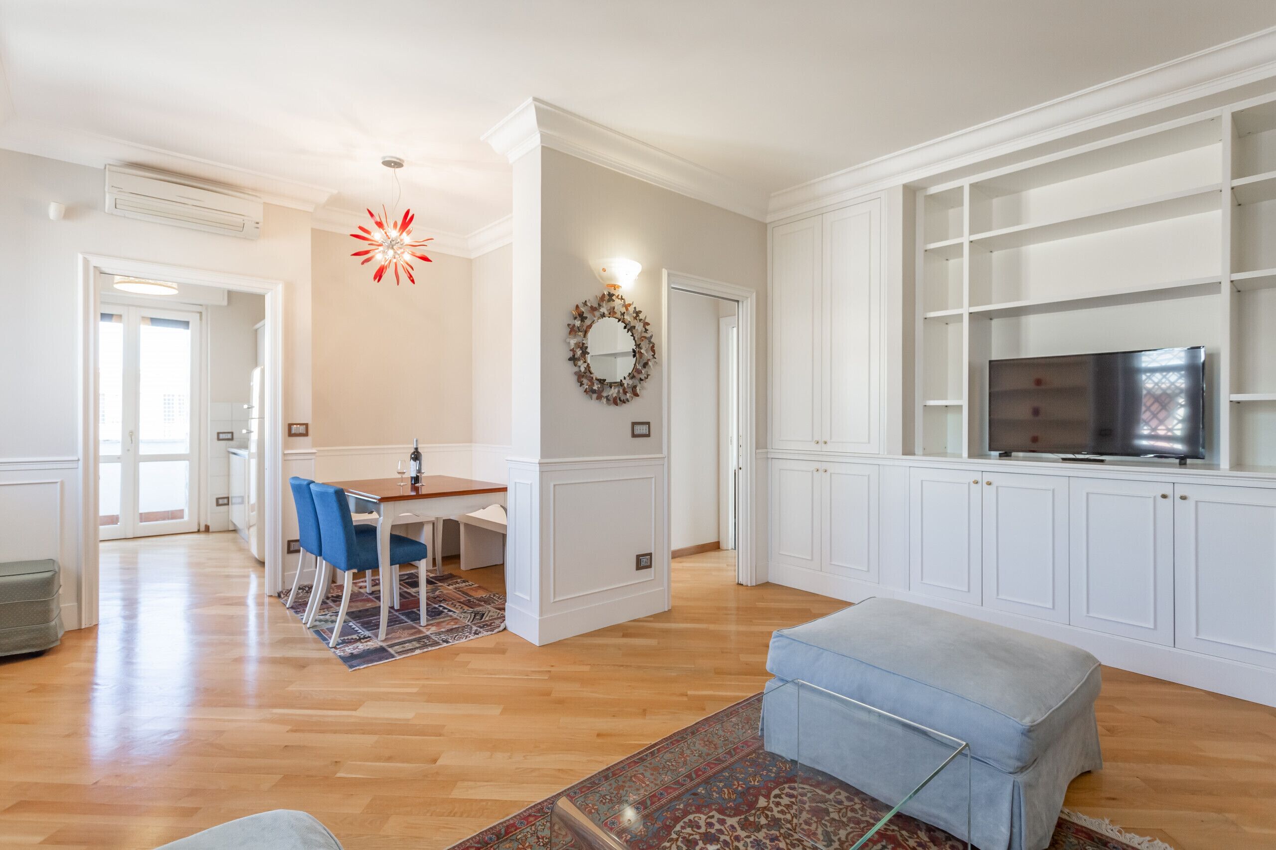 A spacious living room with a wooden floor, a dining area with blue chairs, a light blue ottoman, and a TV on a white cabinet.