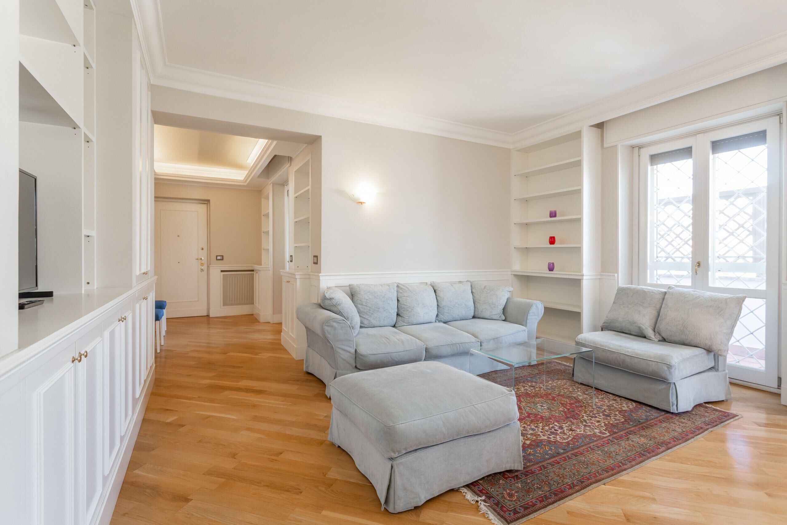 A spacious living room with a light grey sectional sofa, a matching ottoman, and a patterned rug. There are built-in shelves and a television on the left.