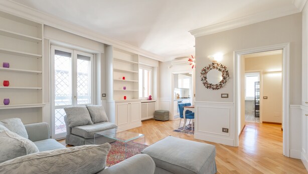 A spacious living room with a grey sofa set, a glass coffee table, and a red patterned rug. There are white built-in shelves and a dining area visible in the background. - Monteverde 2BDR  Penthouse (Roma)