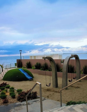 A beachside area with a playground, including a slide and swings, and a view of the ocean.