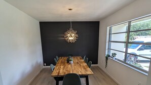 A dining area with a wooden table, six chairs, a chandelier, a potted plant, and a window with a view of a parked car.