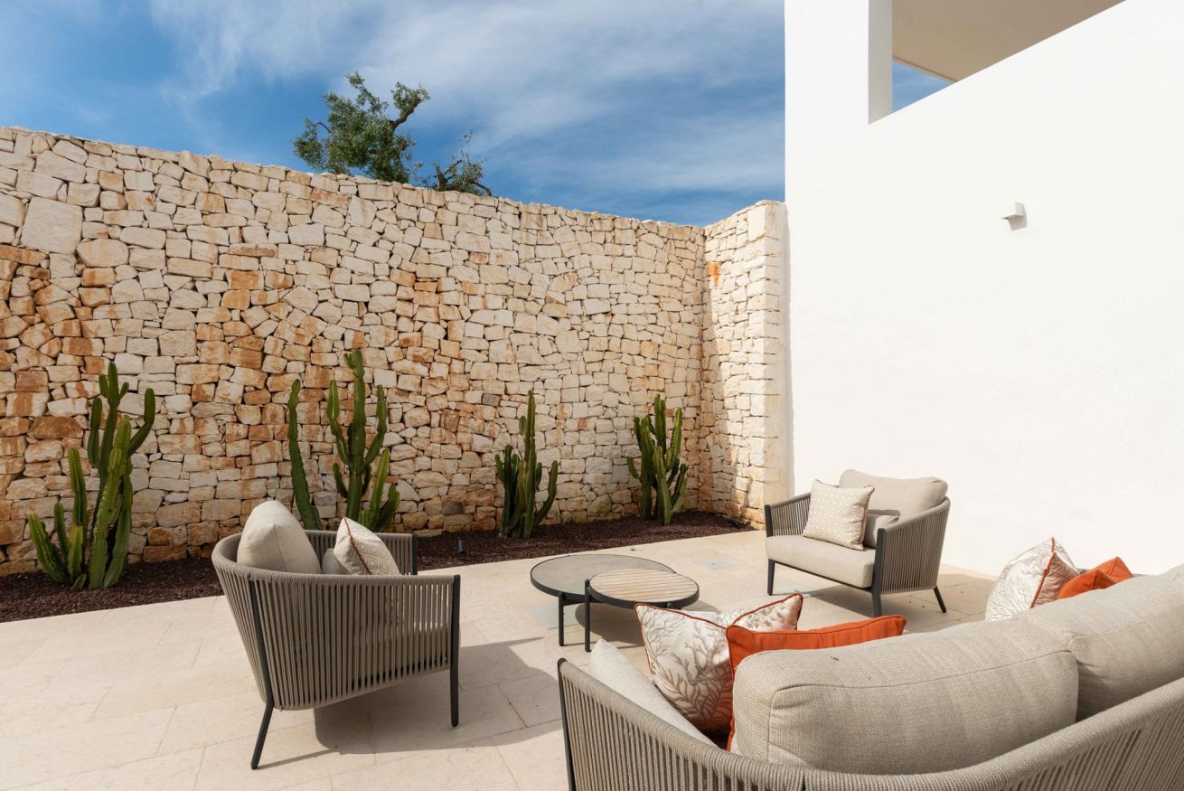 Une terrasse extérieure avec un mur en pierre, des cactus, des meubles en osier et un ciel dégagé.