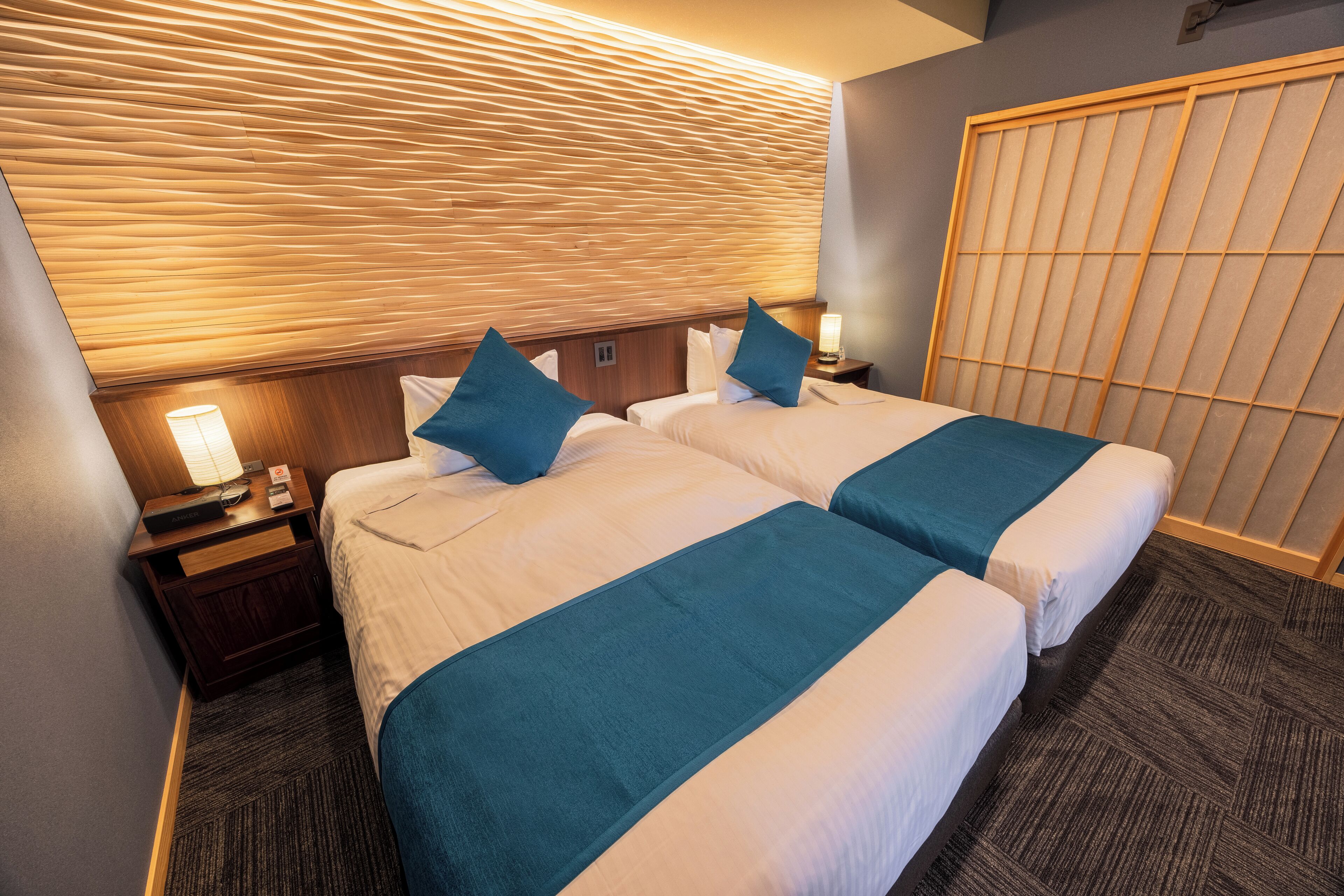 A hotel room with two beds, a wooden headboard, a nightstand with a lamp, and a large window with blinds.