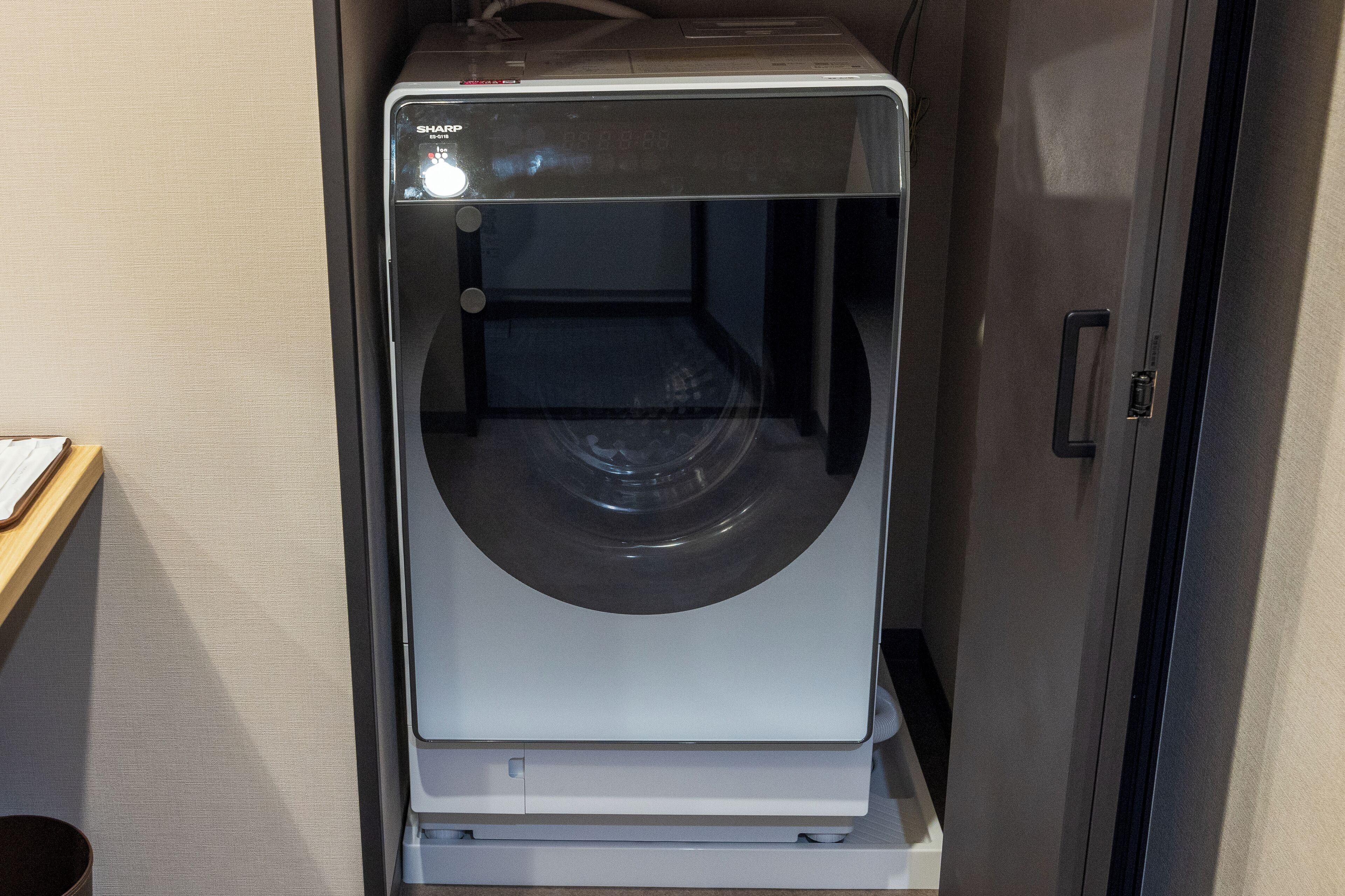 A front view of a compact, modern washing machine with a transparent door, placed in a room with a wooden floor and a beige wall.