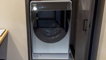 A front view of a compact, modern washing machine with a transparent door, placed in a room with a wooden floor and a beige wall.
