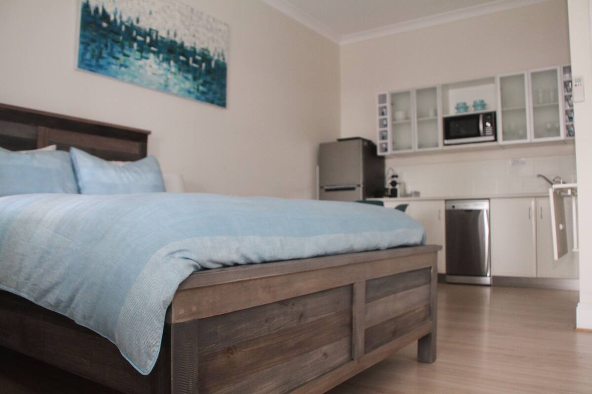 Un dormitorio con cama de madera, mesita de noche y zona de cocina con refrigerador, microondas y lavavajillas.