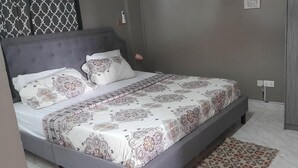 Un dormitorio con una cama tapizada en gris, ropa de cama estampada, una lámpara de noche y un armario.