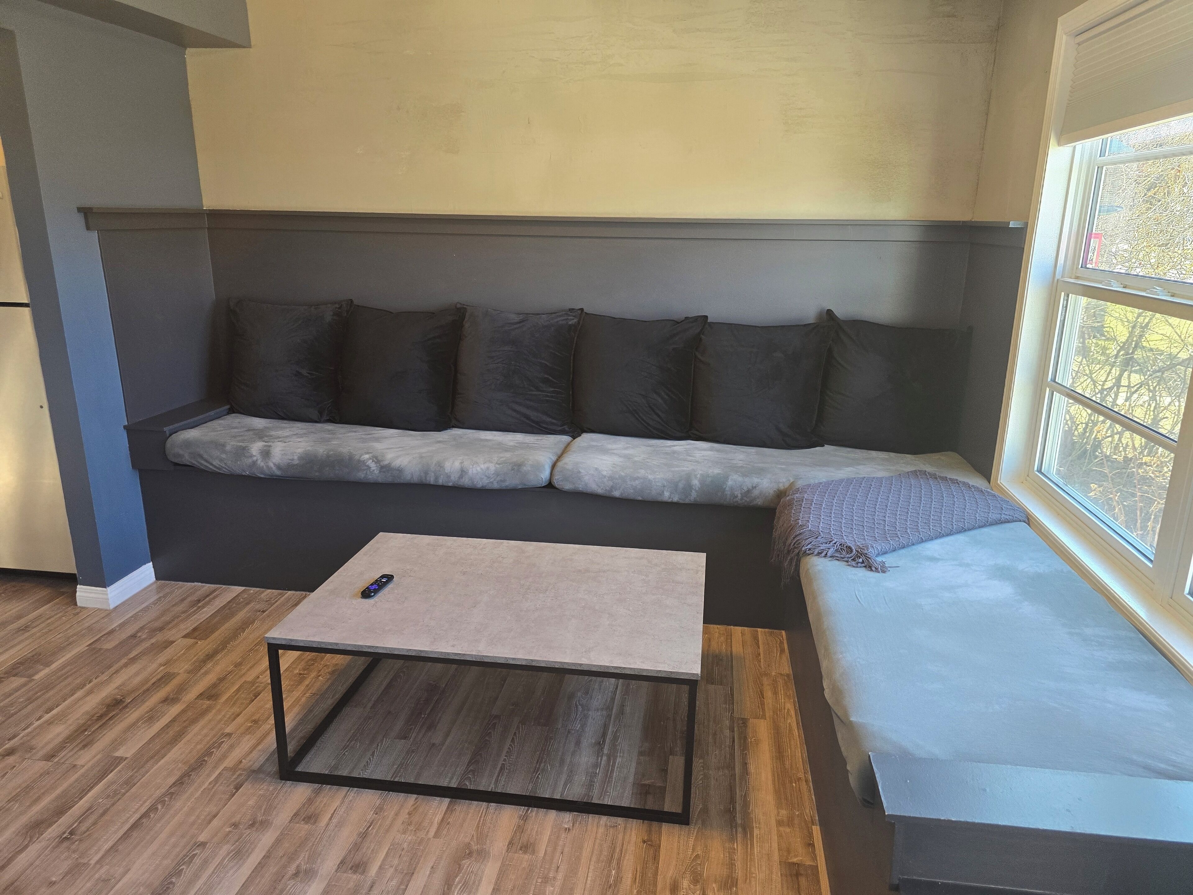 Een moderne woonkamer met een grijze bank, een houten salontafel en een raam met uitzicht op bomen.
