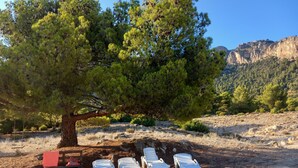 A tree with white plastic lounge chairs under it, a red box, and a rocky hillside in the background. - Cortijo el Corralon Castril, casa 1, casa 2 (Castril)