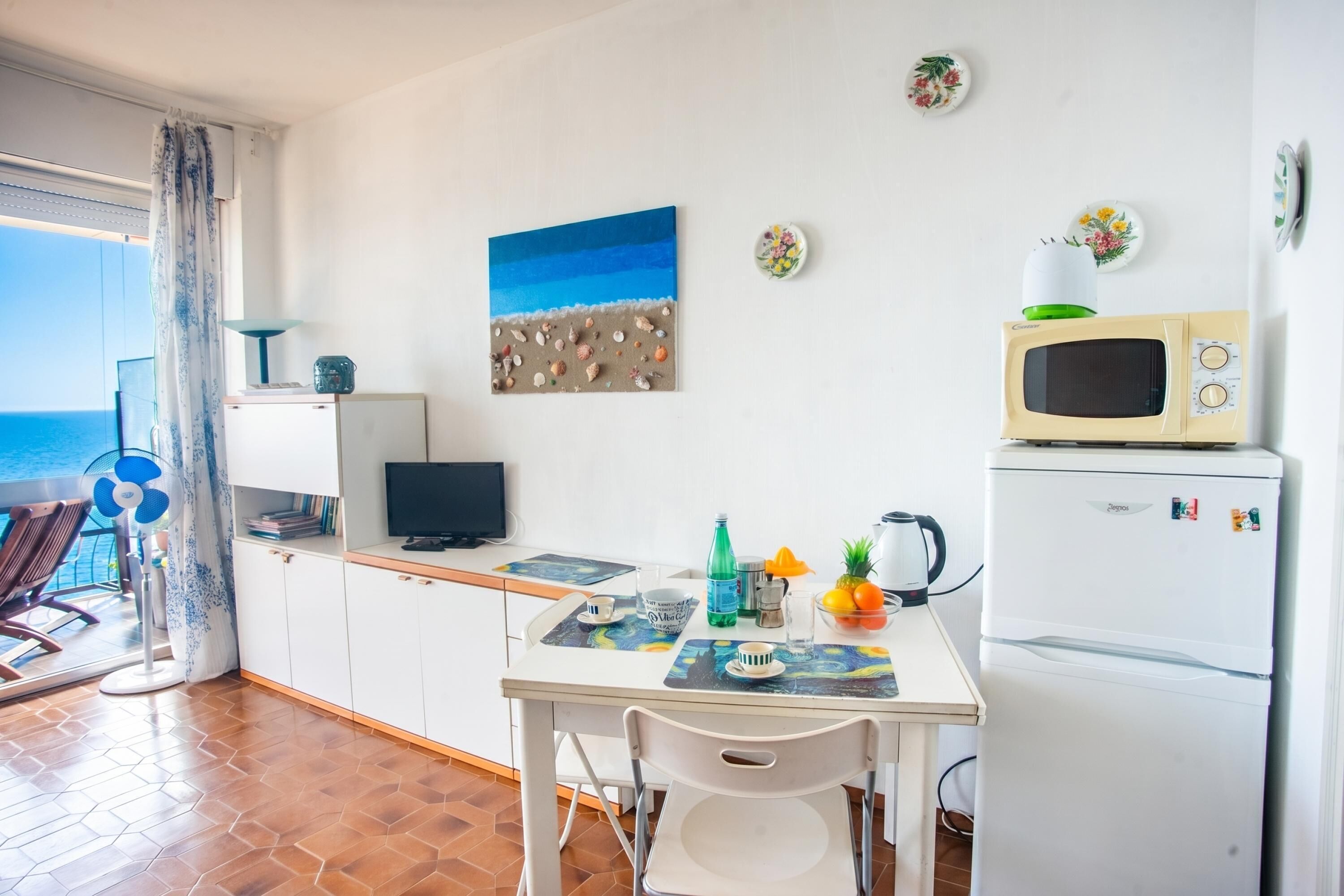 A kitchen with a dining table, chairs, a microwave, a refrigerator, and a beach-themed painting.