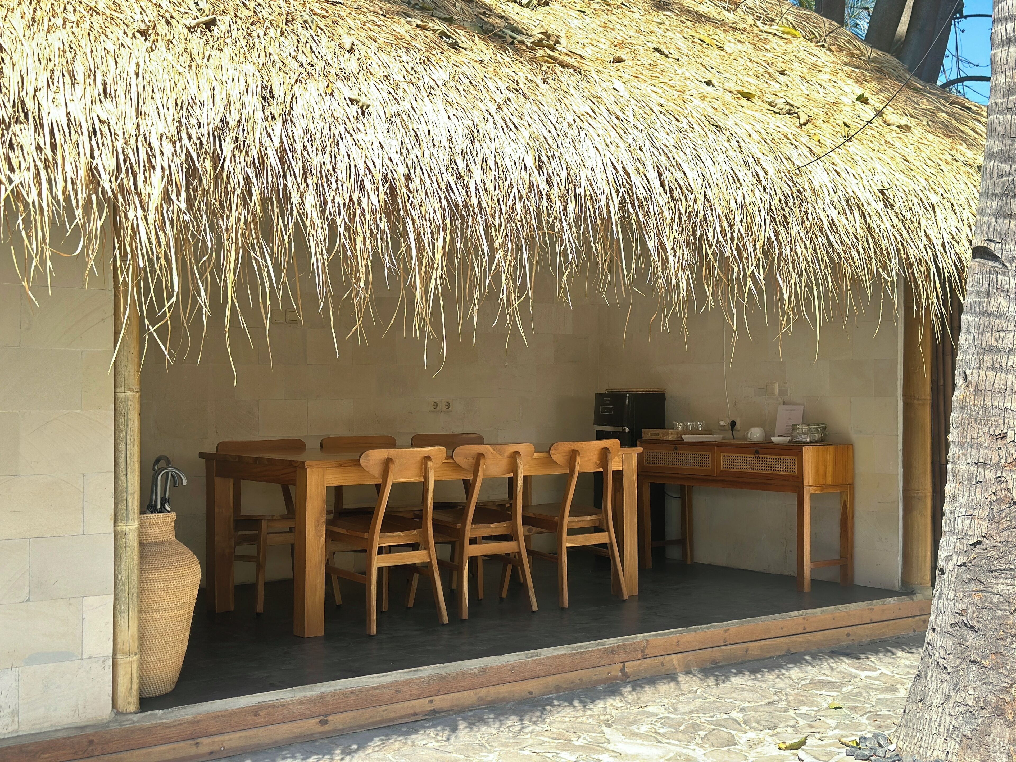 An open-air dining area with a thatched roof, wooden furniture, and a stone floor.