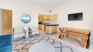 Una sala de estar moderna con un sofá, una mesa de centro redonda y un banco de madera. Se ve una cocina con armarios de madera y vistas al mar a través de una puerta abierta.