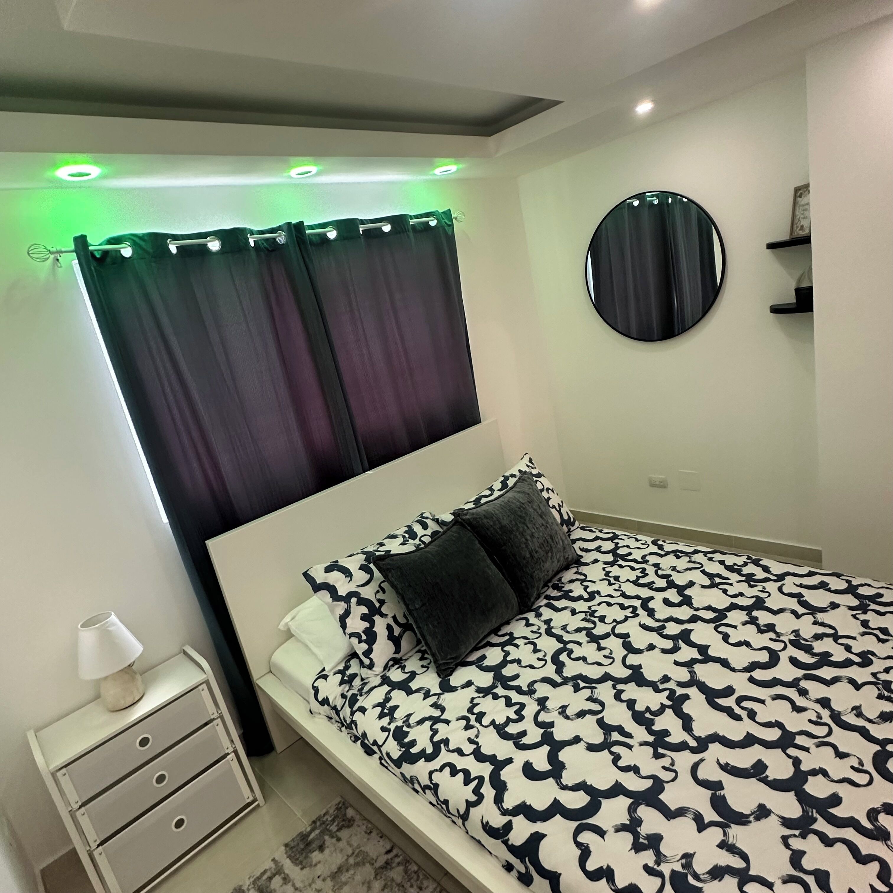 A bedroom with a bed, a bedside table, a lamp, a circular mirror, and a window with curtains.