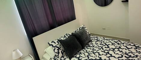 A bedroom with a bed, a bedside table, a lamp, a circular mirror, and a window with curtains.