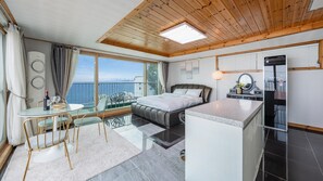 A modern bedroom with a bed, a small sitting area with a table and chairs, a balcony with a view of the sea, and a refrigerator.