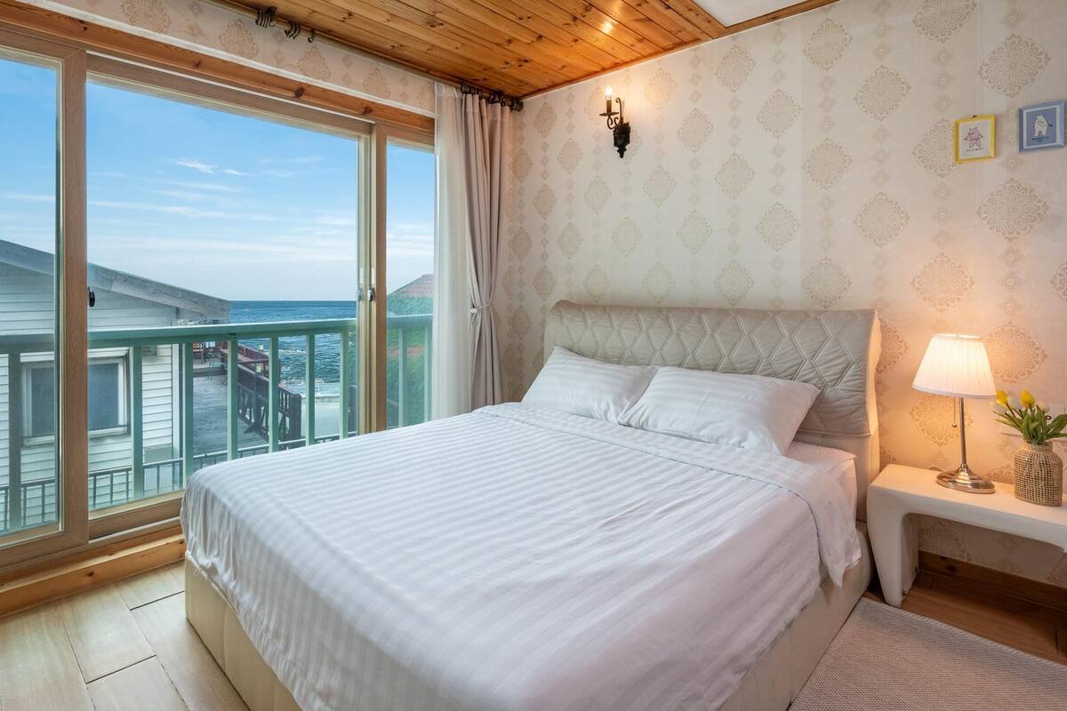 Une chambre avec un grand lit, une table de chevet avec une lampe et une vue sur la mer, visible à travers une porte-fenêtre coulissante.