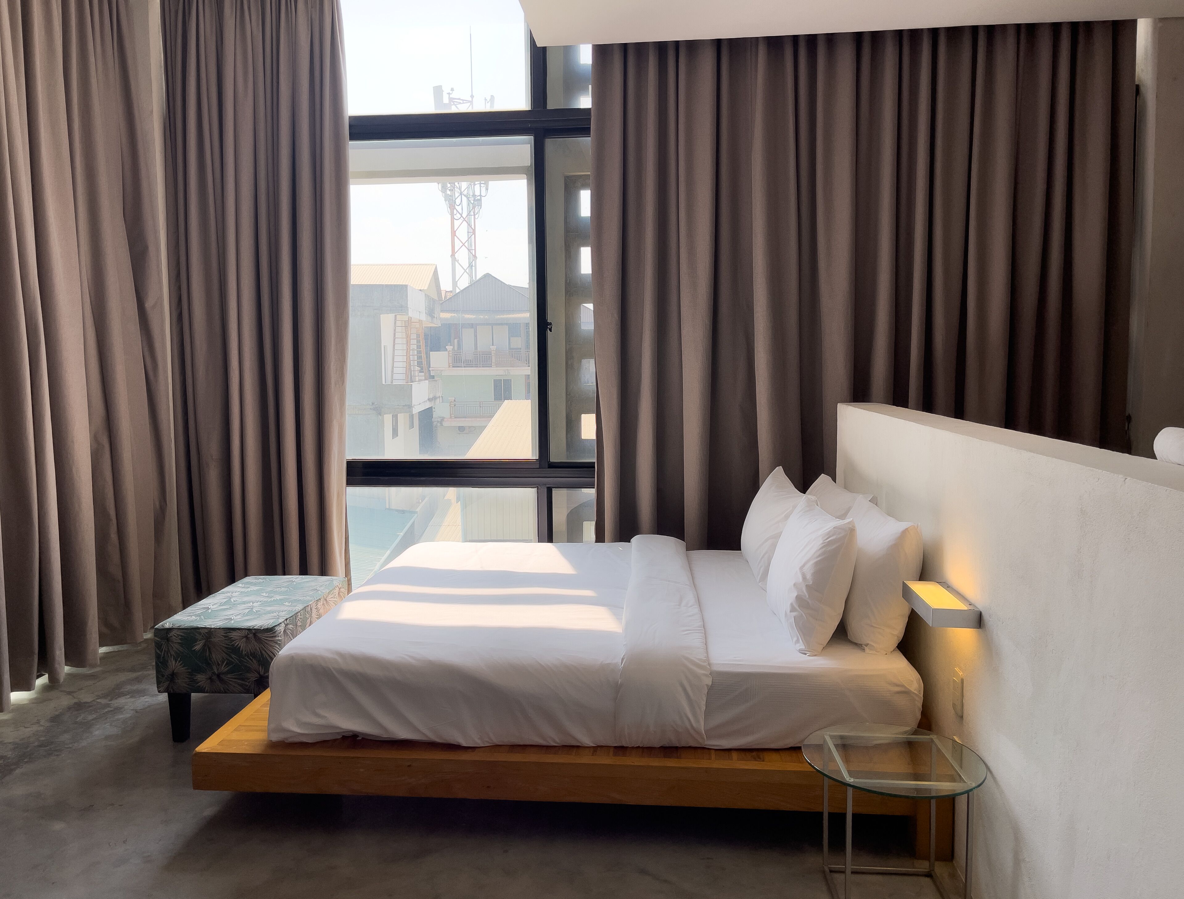 A modern hotel room with a large bed, a glass table, and a view of buildings through the window.
