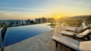 A rooftop infinity pool with lounge chairs and a view of the sunset over the ocean.