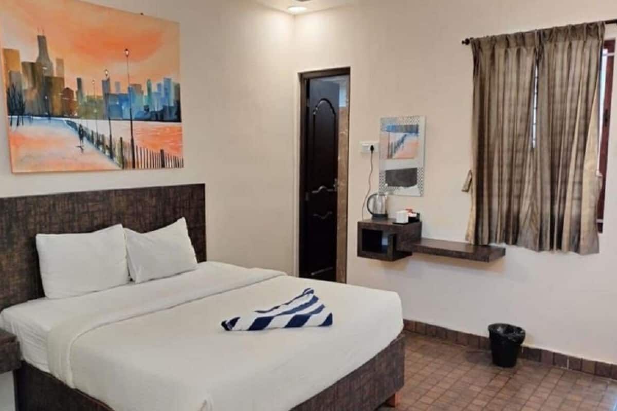 A hotel room with a bed, a nightstand, a painting, a door, a window with curtains, and a small shelf with items.