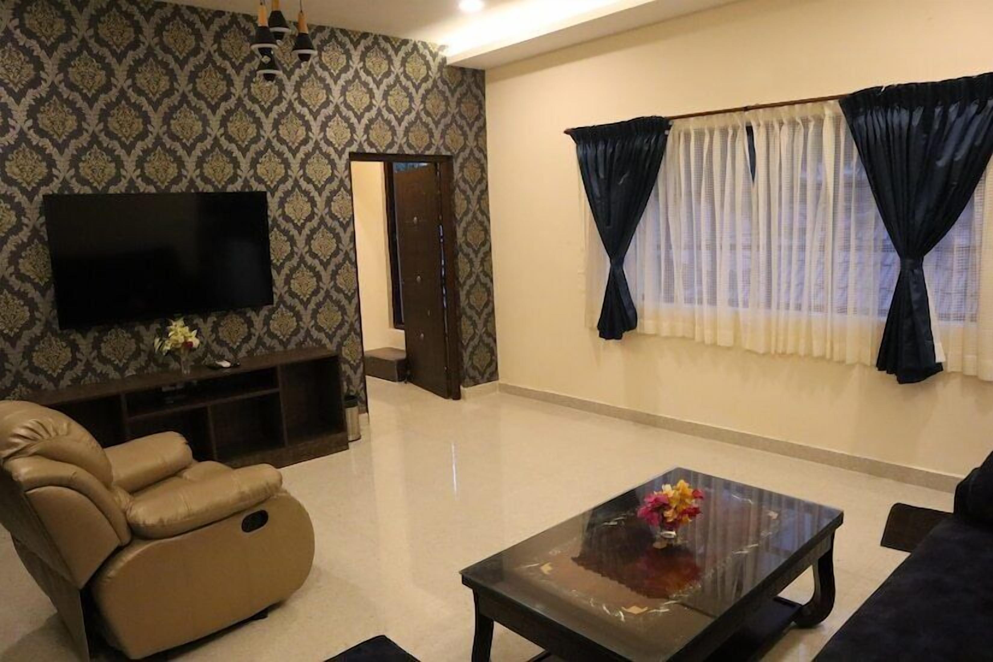 A living room with a flat-screen TV, a reclining chair, a coffee table with flowers, and a patterned wallpaper.