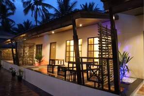 A covered outdoor seating area with tables and chairs, a wooden fence, and a view of palm trees at dusk.