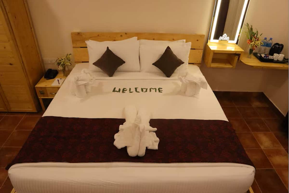 A neatly made bed with a 'WELCOME' sign, a wooden headboard, and a bedside table with a lamp and a vase of flowers.