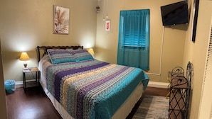 A bedroom with a bed, two bedside lamps, a TV, a window with blue curtains, and a small table with a lamp.