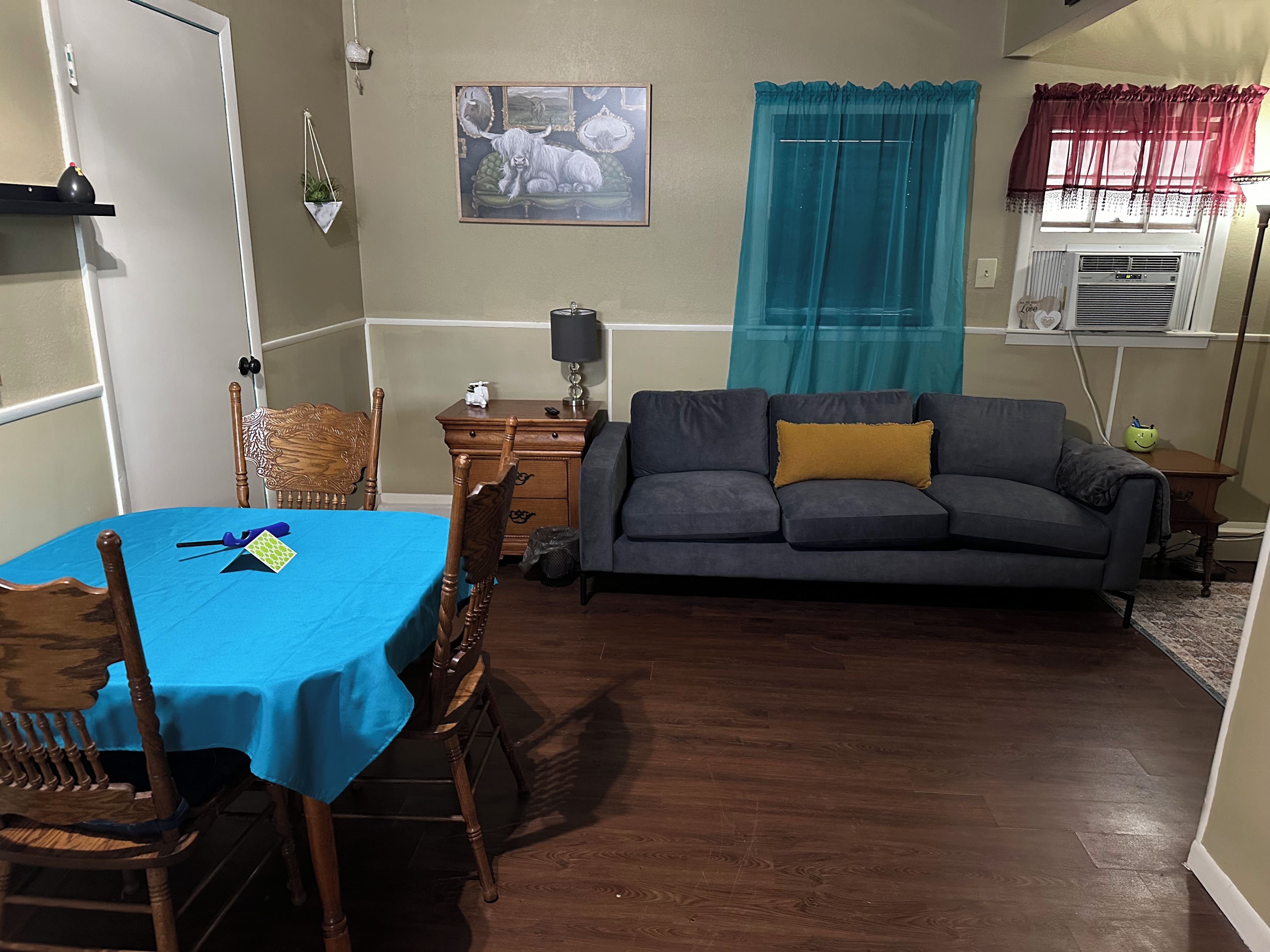 Una habitación con una mesa de comedor, sillas, un sofá, un armario pequeño y una ventana con cortinas.