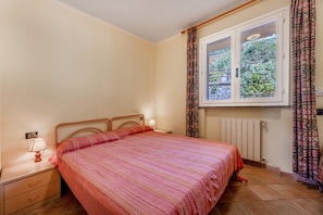 A bedroom with a bed, a nightstand, a lamp, a window with curtains, and a radiator.
