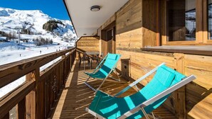 Een houten terras met twee blauwe lounge stoelen, een houten leuning en uitzicht op een besneeuwde berg.