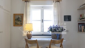 A room with two wooden chairs, a table, a window with white curtains, a lamp, a painting, a basket with flowers, and a bookshelf.