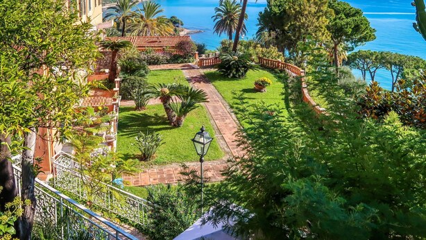 A coastal garden with a pathway leading to a building with arched windows. - Beautiful apartment for 4 guests with WIFI, pool, TV, terrace and panoramic view (Ventimiglia)
