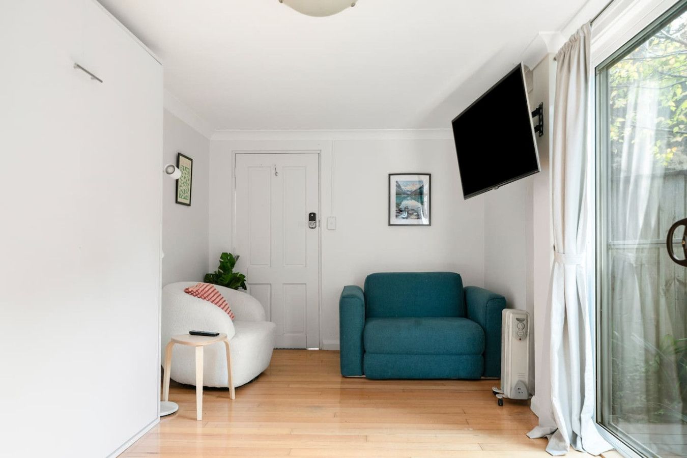 A modern living room with a white sofa, a teal armchair, and a small white table with a remote control. A framed picture hangs on the wall, and a flat-screen TV is mounted above the armchair.