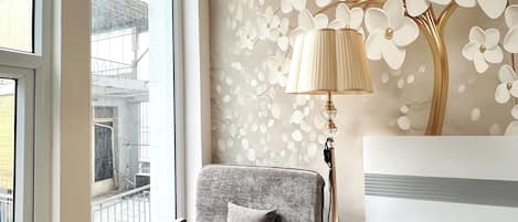 A bedroom with a large window, a chair, a bedside lamp, and a bed.