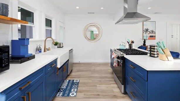 A modern kitchen with blue cabinets, white countertops, and a central island. There is a stove, sink, and a decorative mirror on the wall.