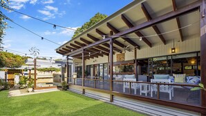 Un espacio al aire libre cubierto con mesas y sillas, una terraza de madera y vista al interior a través de amplios ventanales.