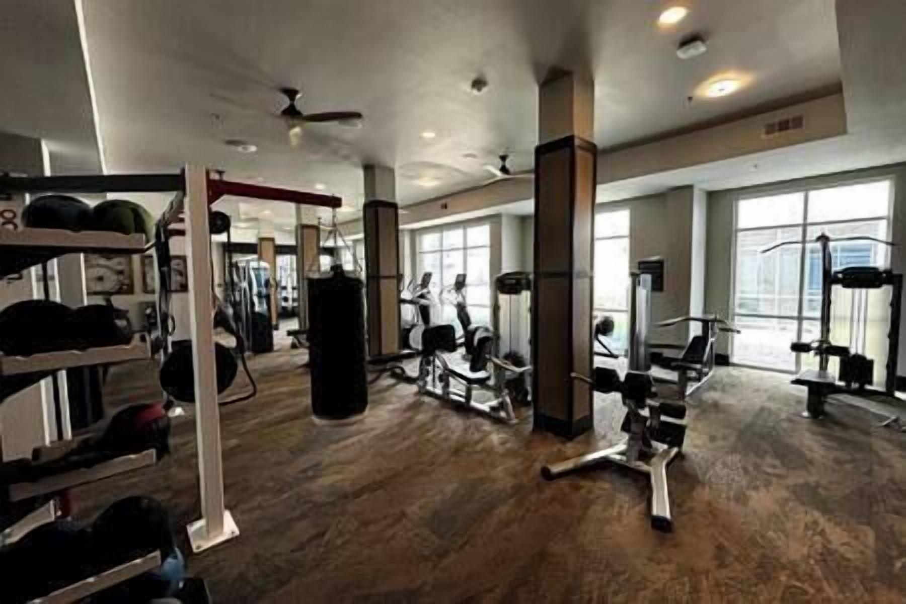 Un gimnasio con diversos equipos de ejercicio, ventanas amplias y suelo de madera.