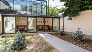 Um edifício moderno com grandes janelas de vidro, área de estar com mesas e cadeiras, e um jardim ajardinado com pequenas árvores.