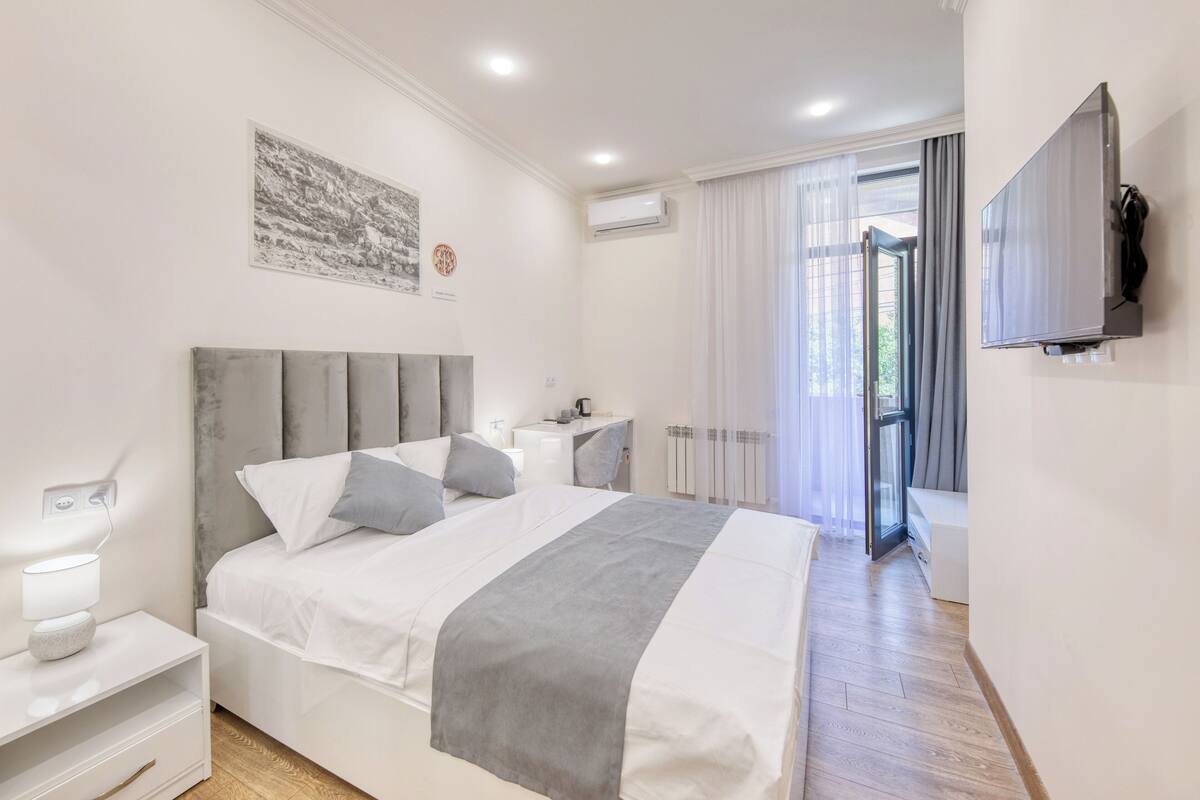 Un dormitorio moderno con cama, mesitas de noche, televisor y ventana con cortinas.