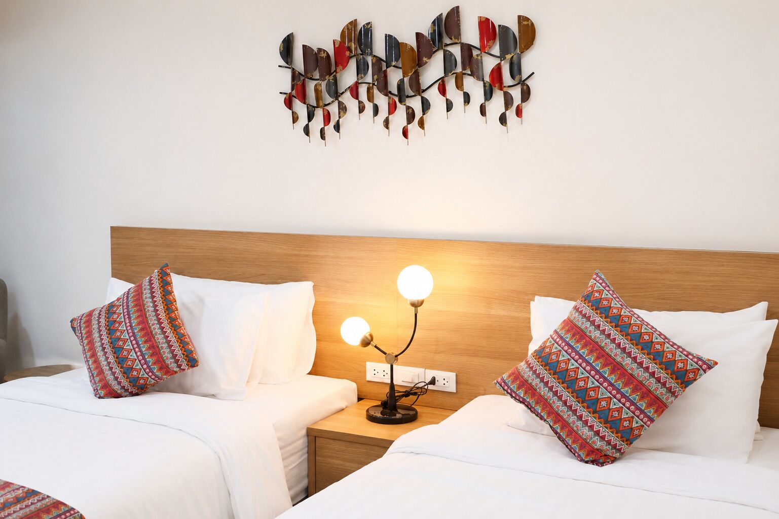 Habitación de hotel con dos camas, ropa blanca, almohadas estampadas, una lámpara fijada a la pared y un cabecero de madera.