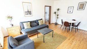 Una sala de estar moderna con un sofá gris, un sillón a juego, una mesita de centro y una mesa de comedor con sillas. Hay dos plantas en macetas, un cuadro con la Torre Eiffel y un mueble de madera.