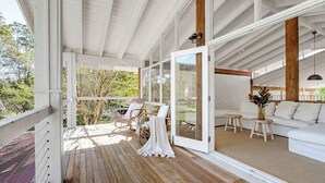 Ein überdachter Außenbereich mit Holzboden, weißen Möbeln und Glastüren zu einem Innenraum mit einem weißen Sofa und einem kleinen Tisch.