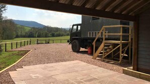 A covered outdoor area with a gravel path leading to a wooden structure, a truck, and a view of green hills. - Unique stay in your very own army truck. Sleeps 4. Hot tub. Tranquil setting. (Llangwm)
