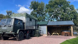 A military-style camper parked next to a wooden cabin with a gravel driveway and a picnic table. - Unique stay in your very own army truck. Sleeps 4. Hot tub. Tranquil setting. (Llangwm)