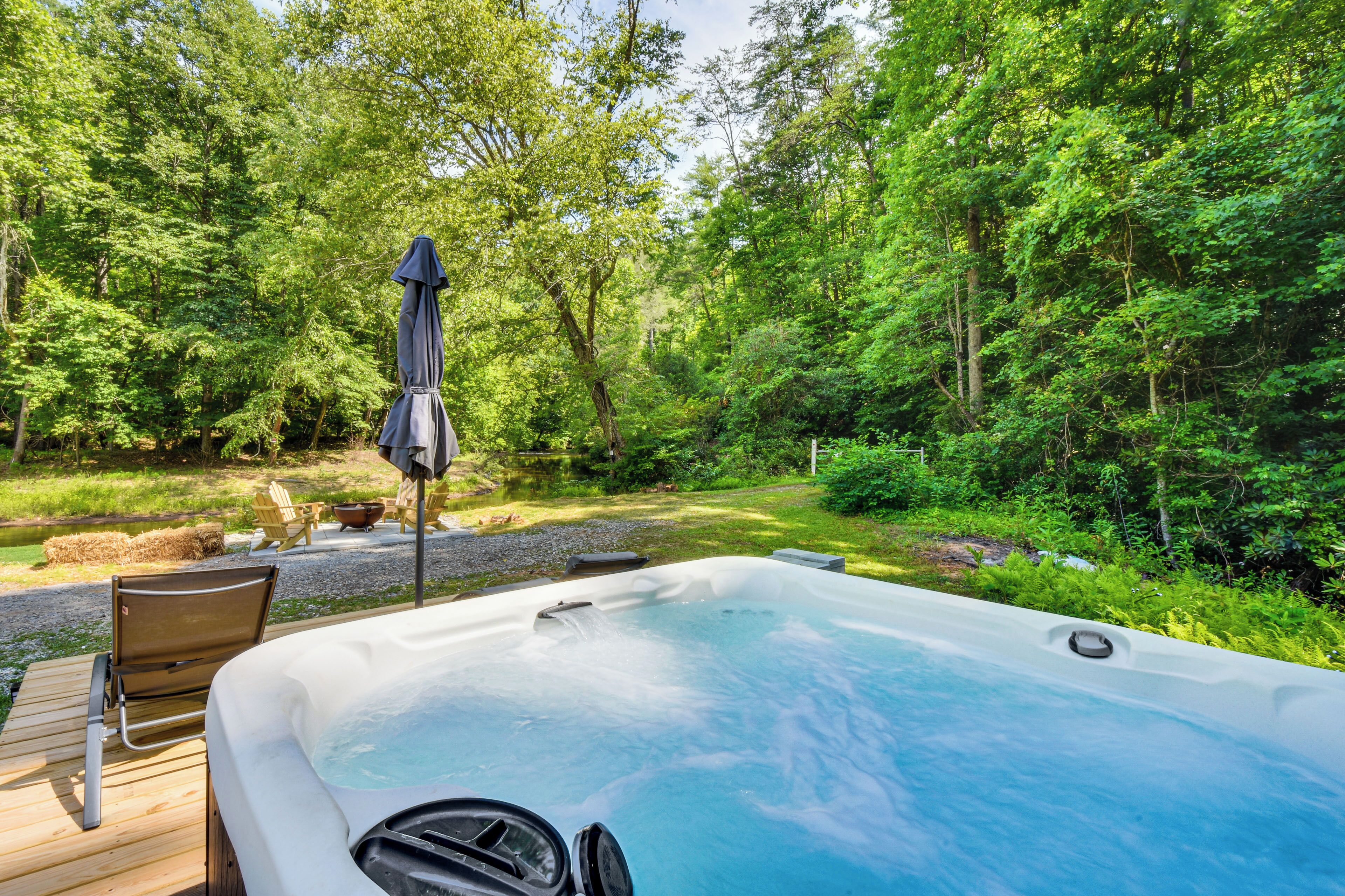 A hot tub in a forested area with a wooden deck, chairs, and a table.