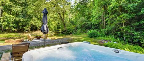 A hot tub in a forested area with a wooden deck, chairs, and a table.