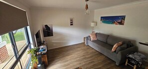 Ein Wohnzimmer mit grauem Sofa, einem kleinen Tisch mit Lampe, einem Fernseher und einem Fenster mit Jalousien.