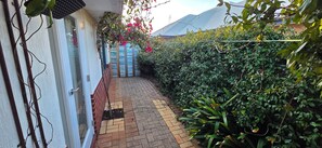 Ein schmaler Ziegelweg führt zu einem Hauseingang, umgeben von dichtem Grün und einer Kletterpflanze mit rosa Blüten.